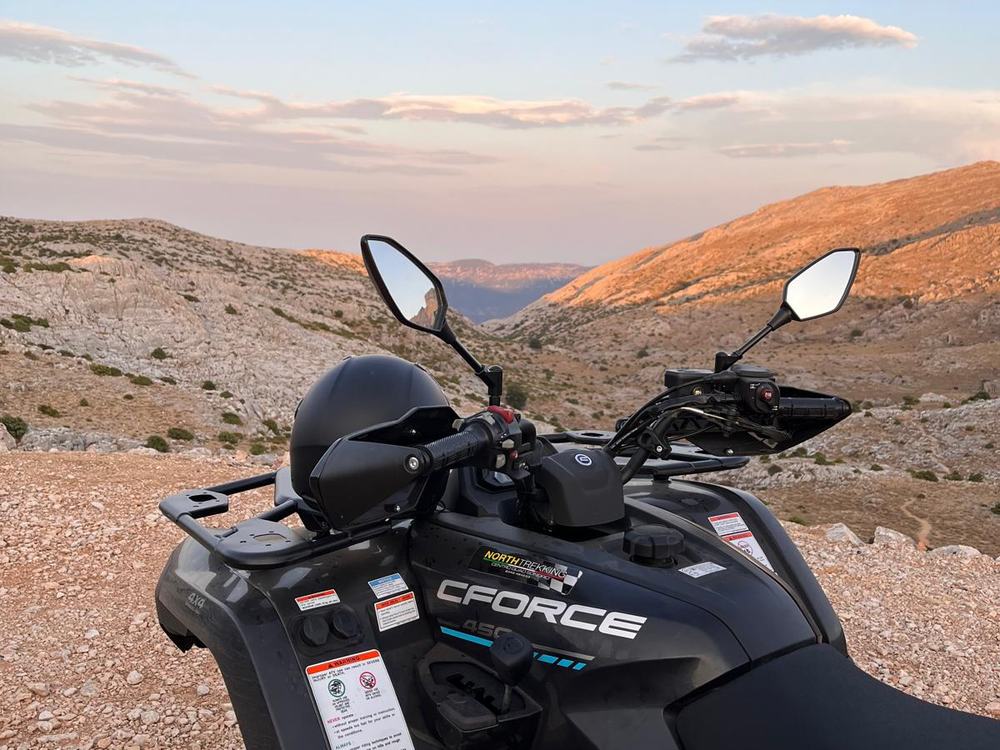 Quad nero CFORCE 450 con casco sul portapacchi, fermo su sterrato davanti a una valle montuosa al tramonto